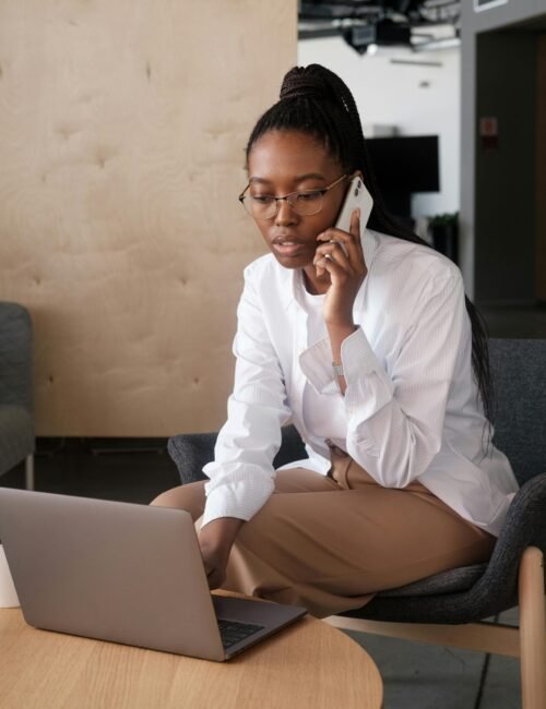 African American businesswoman talking on smartphone while working on laptop indoors.